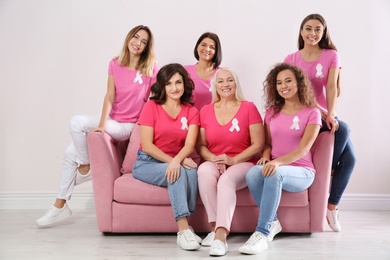 Group of women with silk ribbons sitting on sofa against light wall. Breast cancer awareness concept