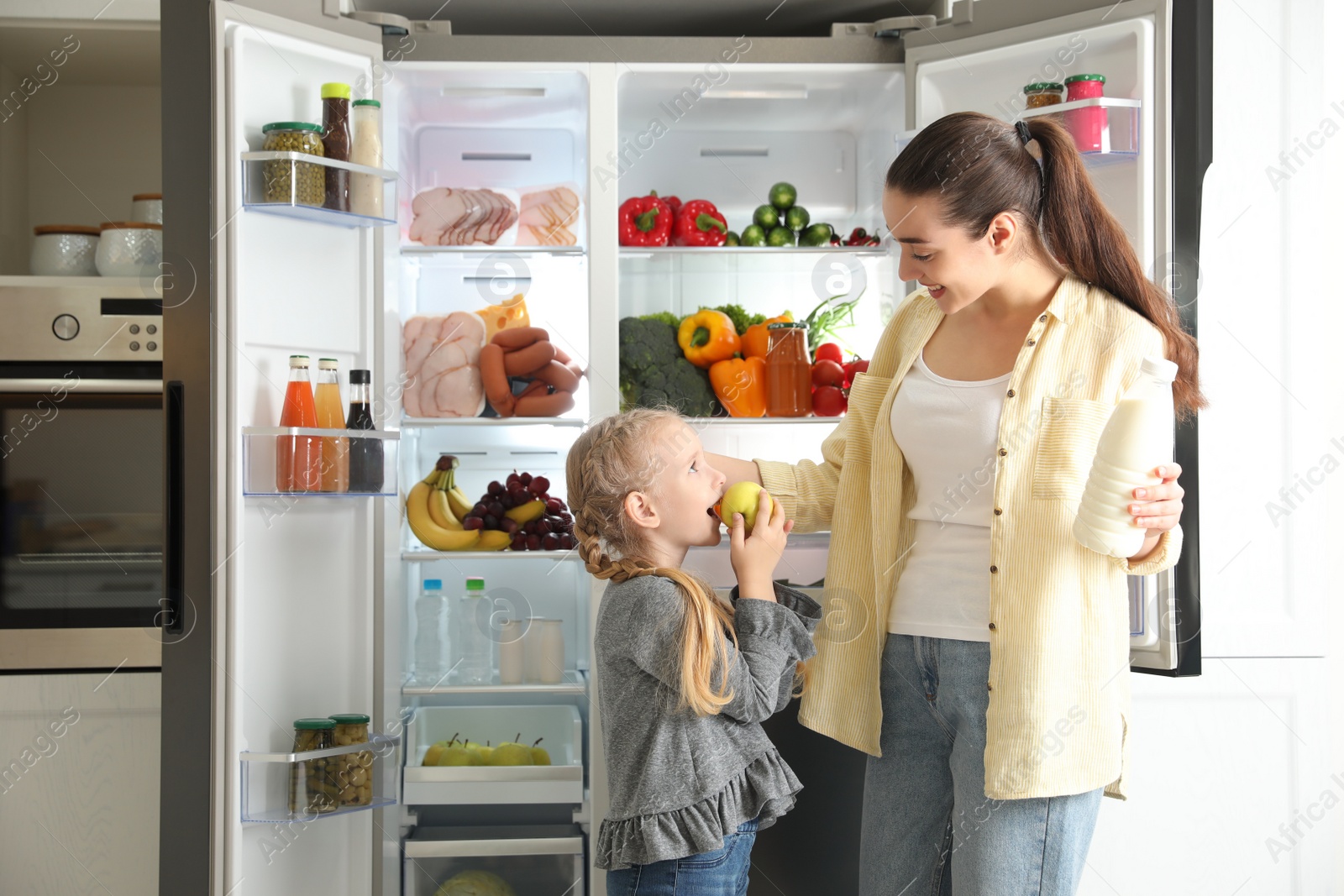 Photo of Young mother and her daughter with products near open refrigerator in kitchen