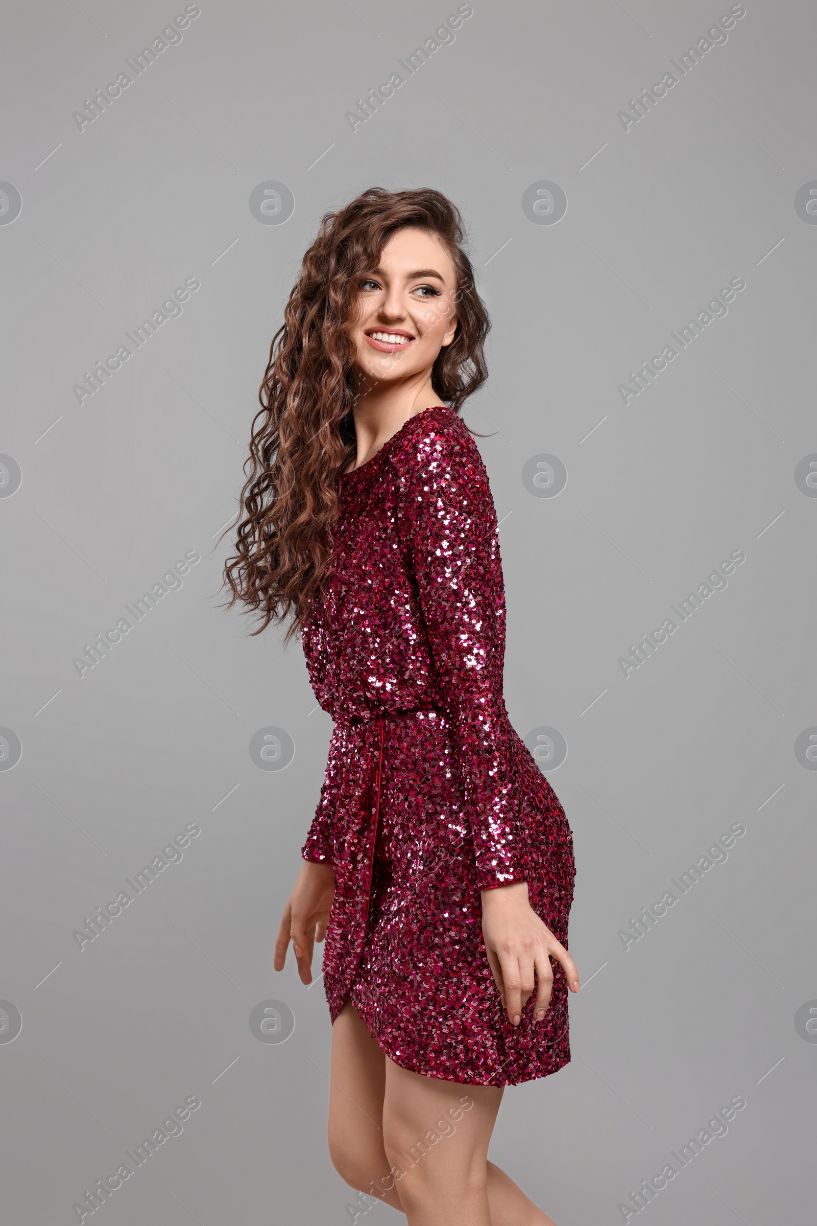 Photo of Beautiful young woman with long curly brown hair in pink sequin dress on grey background