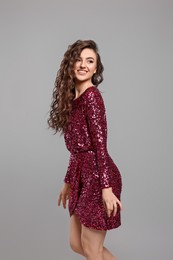 Beautiful young woman with long curly brown hair in pink sequin dress on grey background