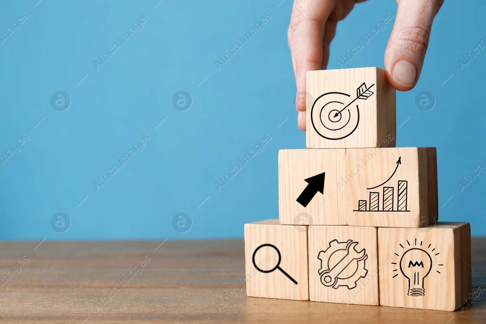 Image of Woman building pyramid of cubes with icons on wooden table against light blue background, closeup. Target, arrow, magnifier and other illustrations