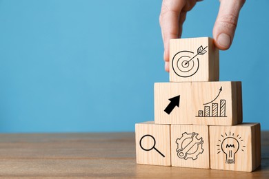 Woman building pyramid of cubes with icons on wooden table against light blue background, closeup. Target, arrow, magnifier and other illustrations