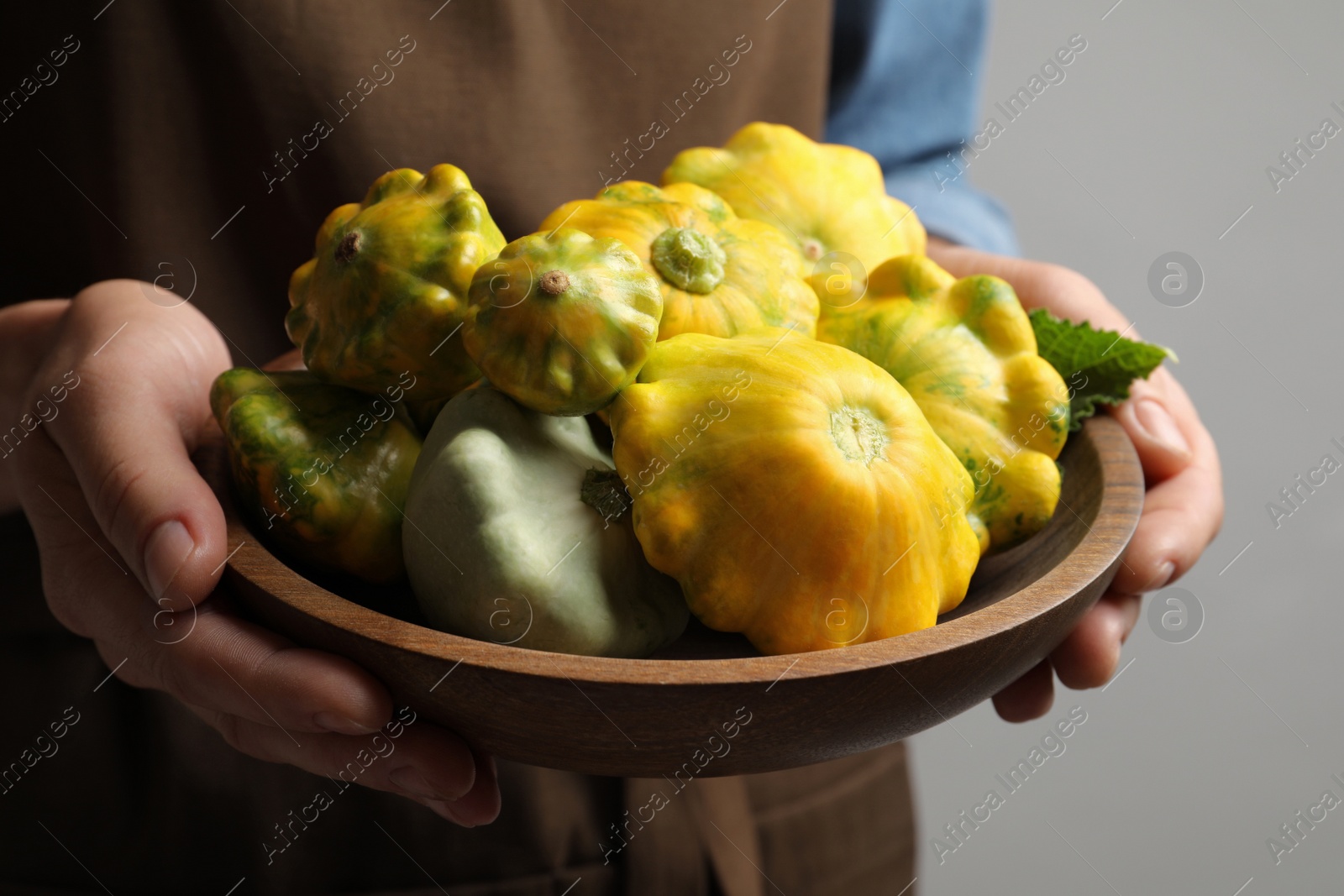 Photo of Woman with wooden bowl of fresh ripe pattypan squashes on grey background, closeup