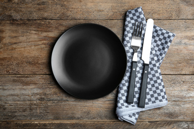 Photo of Empty black plate with cutlery and napkin on wooden table, flat lay