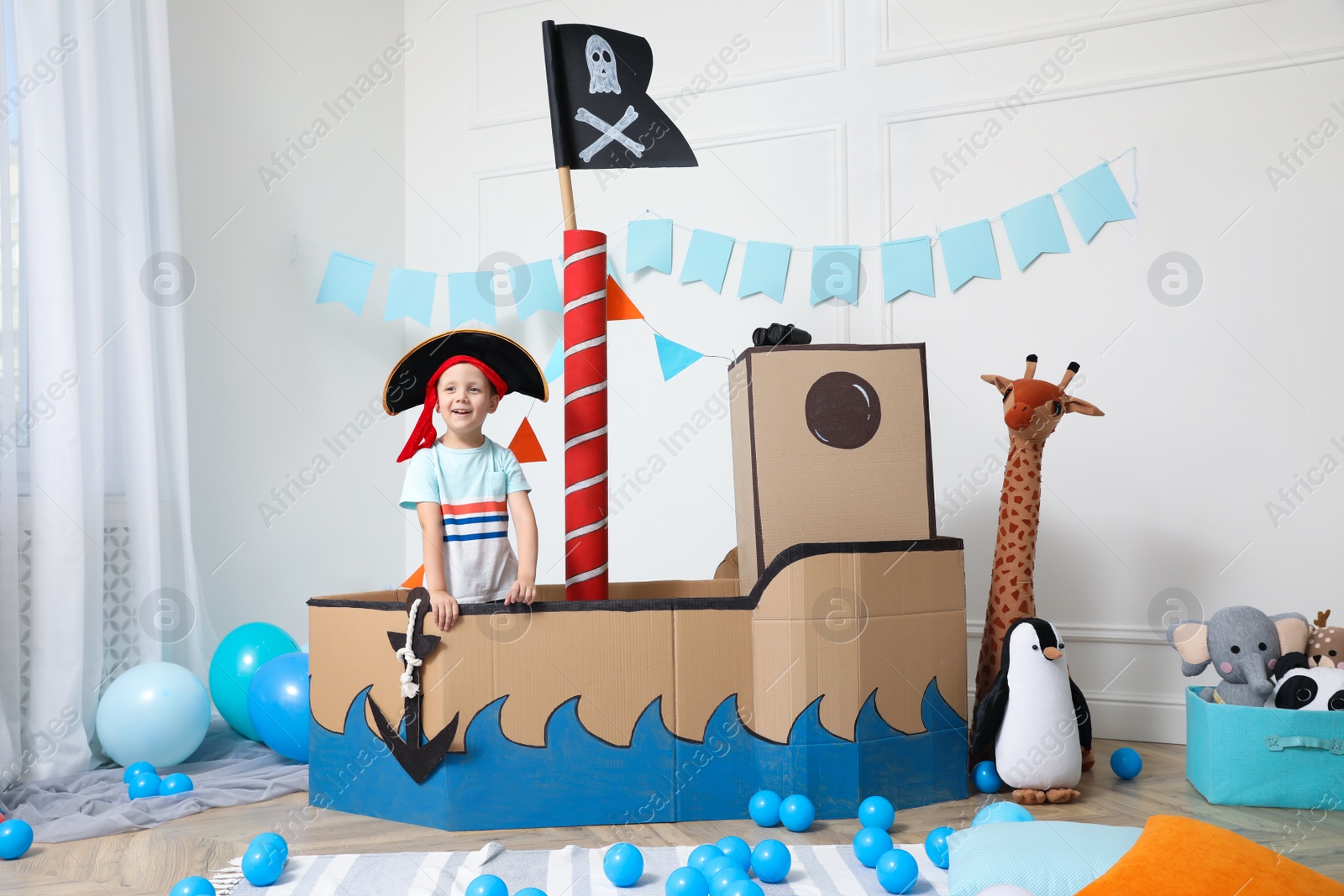 Photo of Cute little boy playing with pirate cardboard ship and toys at home. Child's room interior