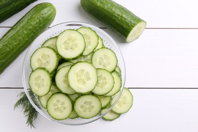 Photo of Cut cucumber in glass bowl, fresh vegetables and dill on white wooden table, flat lay. Space for text