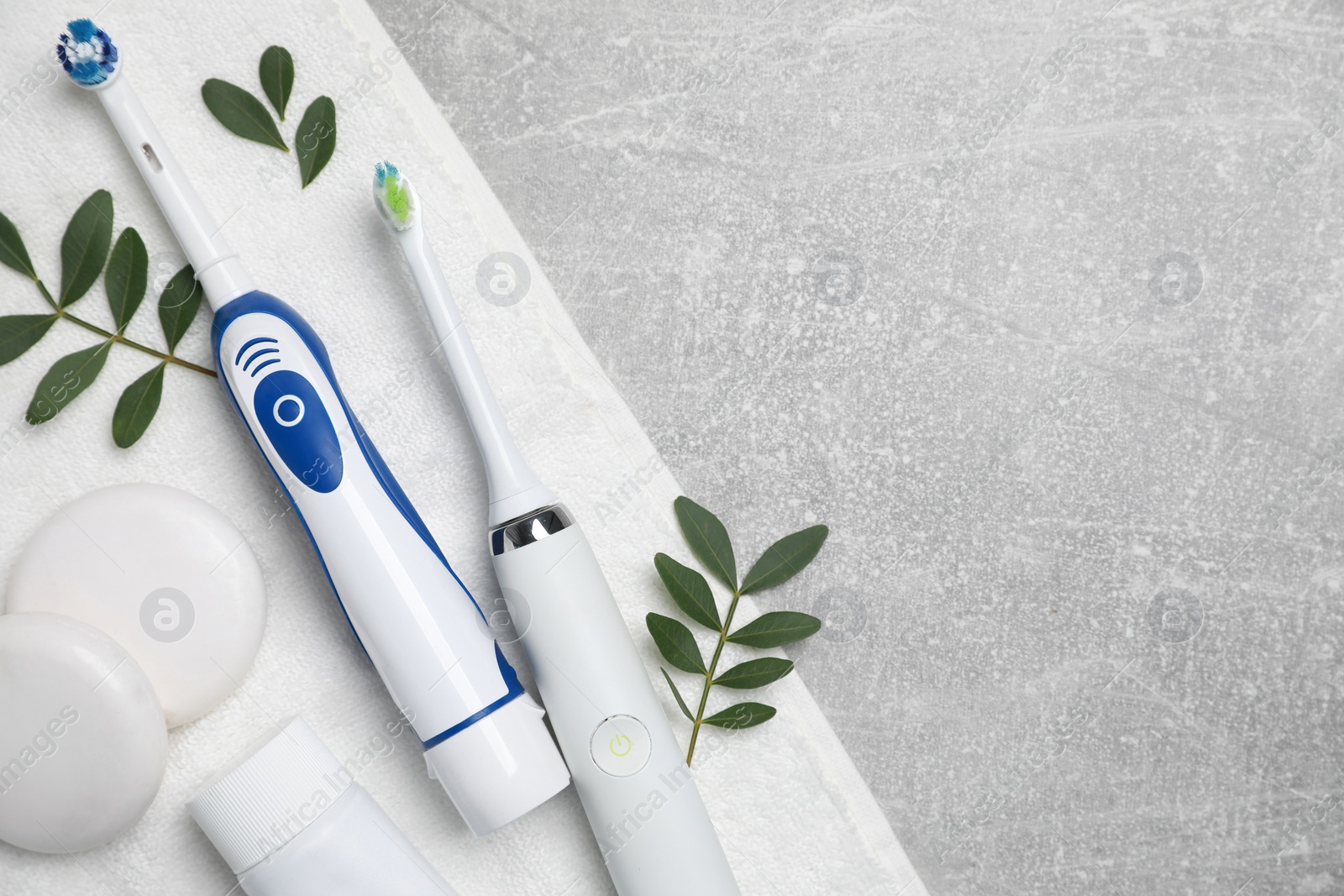 Photo of Electric toothbrushes, towel, tube of paste and leaves on grey textured table, top view. Space for text