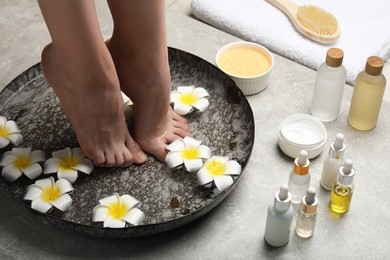 Woman soaking her feet in bowl with water and flowers on light grey floor, closeup. Spa treatment