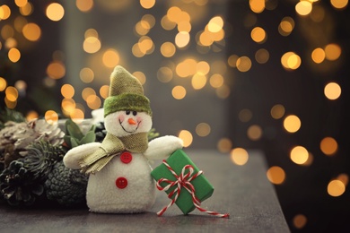 Image of Cute snowman with gift and Christmas decor on table, bokeh effect. Space for text