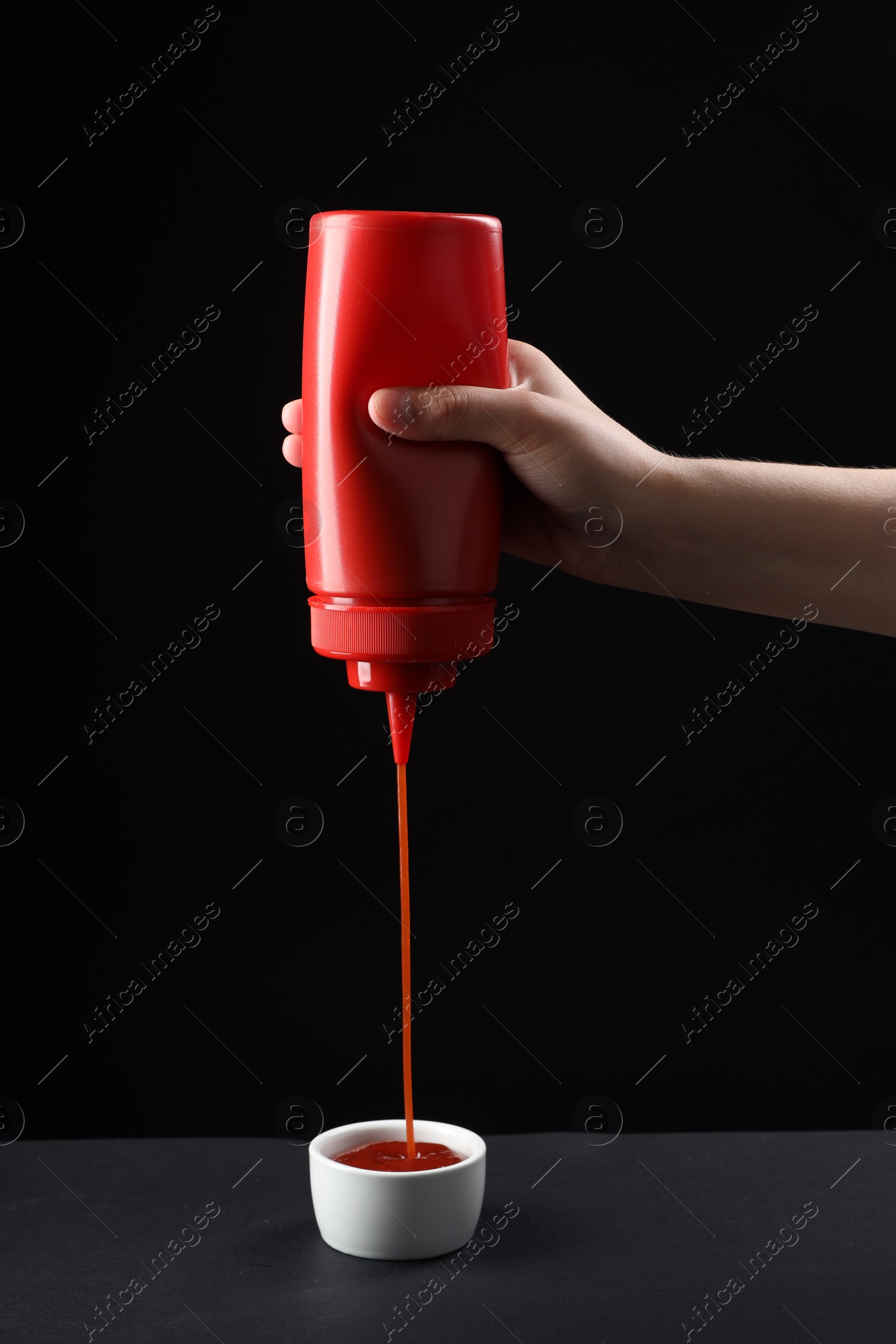 Photo of Woman pouring tasty ketchup from bottle into bowl on table against black background, closeup