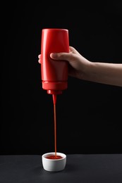 Woman pouring tasty ketchup from bottle into bowl on table against black background, closeup