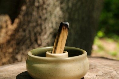 Photo of Smoldering palo santo stick in holder on wooden stump outdoors, closeup