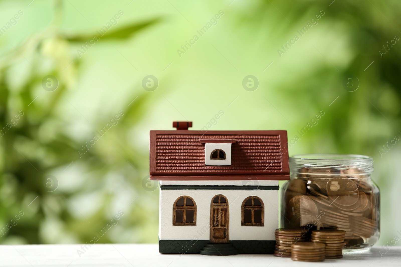 Photo of Model of house and jar with coins on table against blurred green background. Space for text