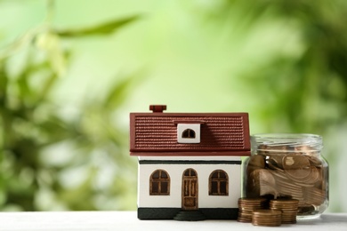 Photo of Model of house and jar with coins on table against blurred green background. Space for text