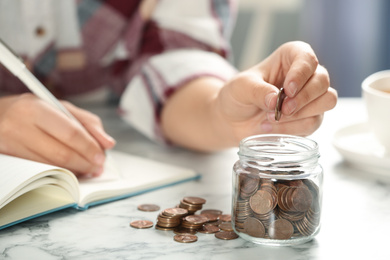 Photo of Woman putting money into glass jar at white marble table, closeup