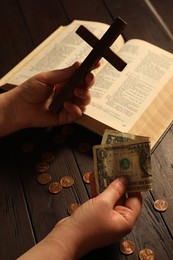 Donate and give concept. Woman holding dollar banknote and cross at wooden table, closeup