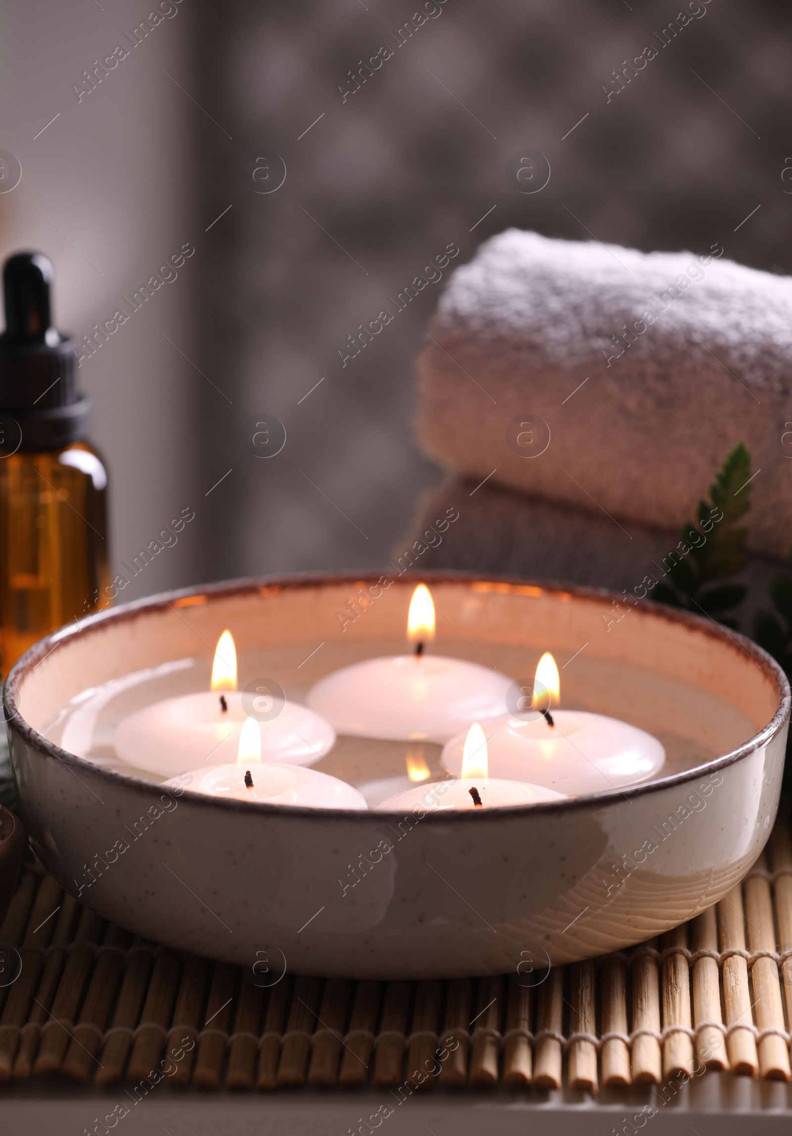 Photo of Spa composition. Burning candles in bowl, towels and bottle of essential oil on table, closeup