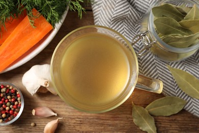 Hot delicious bouillon in glass cup and ingredients on wooden table, flat lay