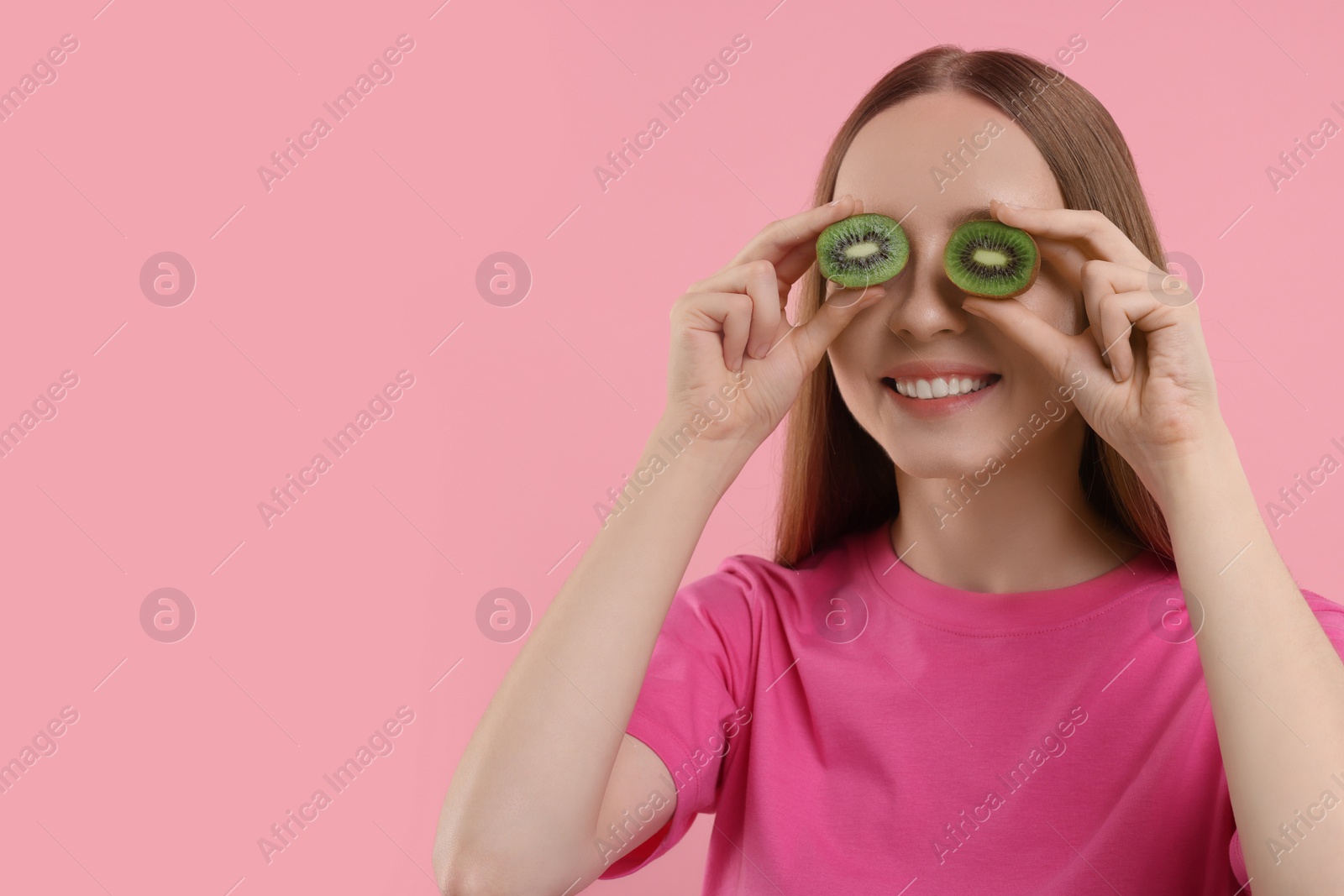 Photo of Young woman holding halves of kiwi near her eyes on pink background. Space for text