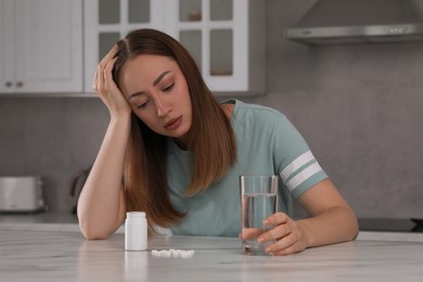 Photo of Depressed woman with glass of water and antidepressant pills at table in kitchen