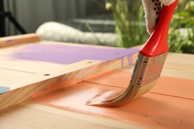 Photo of Worker applying coral paint onto wooden surface against blurred background, closeup. Space for text