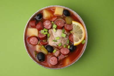 Photo of Meat solyanka soup with thin dry smoked sausages in bowl on light green background, top view