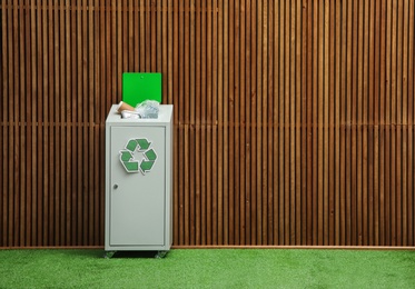 Overfilled trash bin with recycling symbol near wooden wall indoors. Space for text