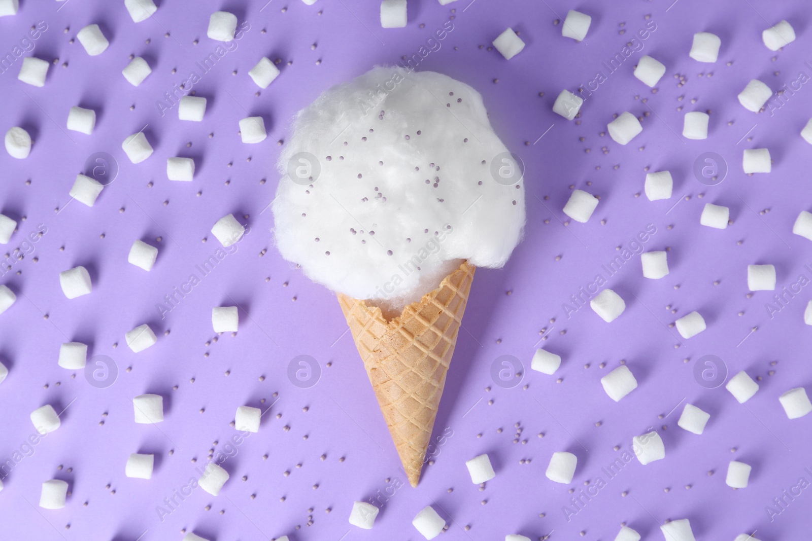 Photo of Sweet cotton candy in waffle cone surrounded by marshmallows on purple background, flat lay