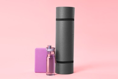 Photo of Grey exercise mat, yoga block and bottle of water on pink background