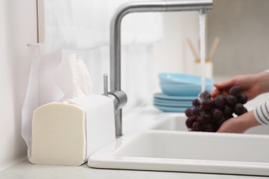 Photo of Woman washing grapes with running water over sink in kitchen, focus on paper towels. Space for text