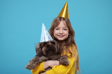 Photo of Happy little girl in party hat hugging dog on light blue background