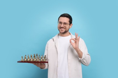 Photo of Smiling man holding chessboard with game pieces and showing OK gesture on light blue background