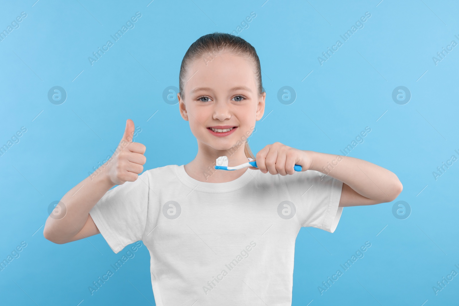 Photo of Happy girl brushing her teeth with toothbrush and showing thumb up on light blue background