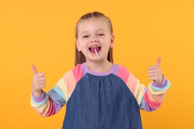 Photo of Happy little girl eating lollipop and showing thumbs up on orange background