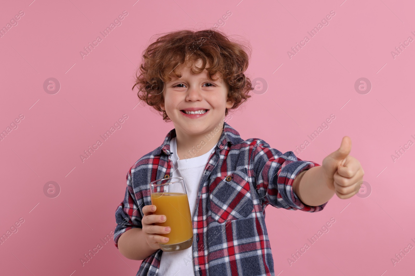 Photo of Cute little boy with glass of fresh juice showing thumb up on pink background