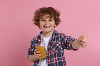 Cute little boy with glass of fresh juice showing thumb up on pink background