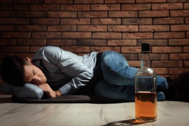 Photo of Addicted man lying near red brick wall, focus on bottle of alcoholic drink