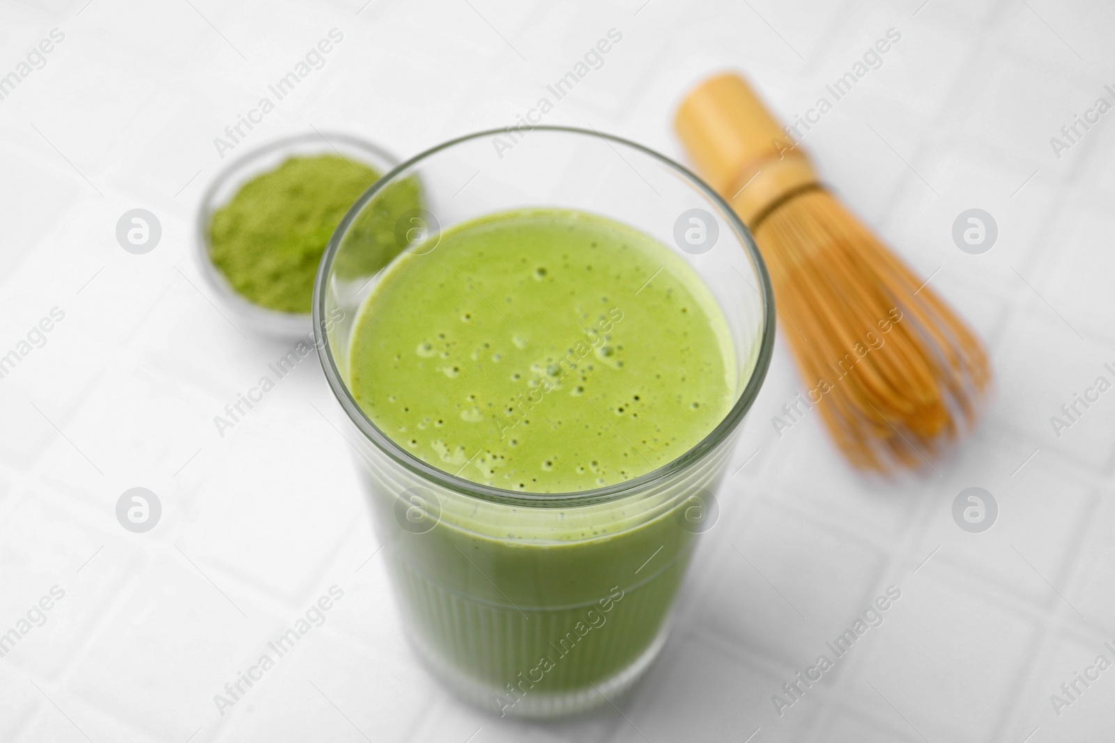 Photo of Glass of tasty matcha smoothie, powder and bamboo whisk on white tiled table, closeup