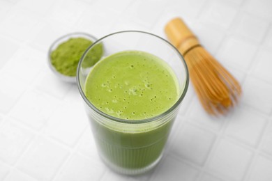 Photo of Glass of tasty matcha smoothie, powder and bamboo whisk on white tiled table, closeup