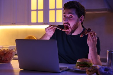Photo of Man eating pizza while using laptop in kitchen at night. Bad habit