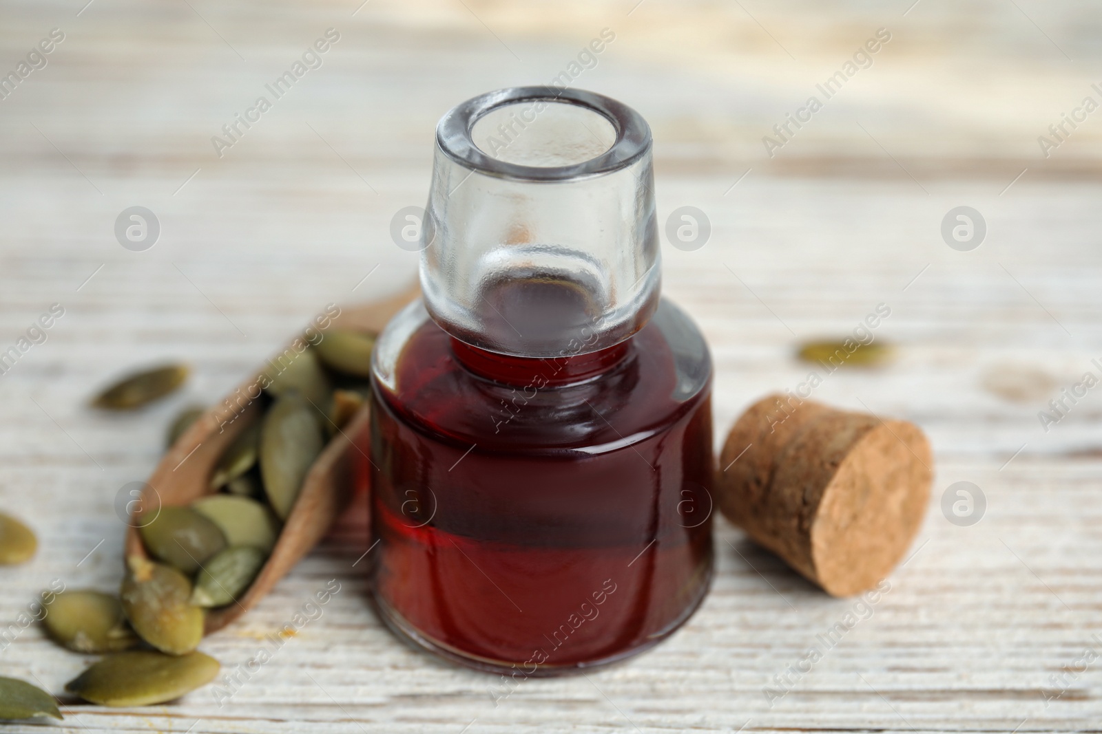 Photo of Glass bottle of oil and scoop with pumpkin seeds on white wooden table, closeup