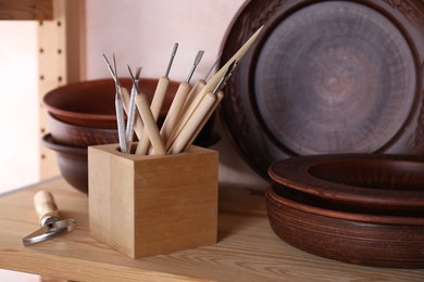 Photo of Set of different crafting tools and clay dishes on wooden rack in workshop