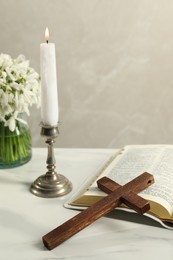 Burning church candle, wooden cross, Bible and flowers on white marble table