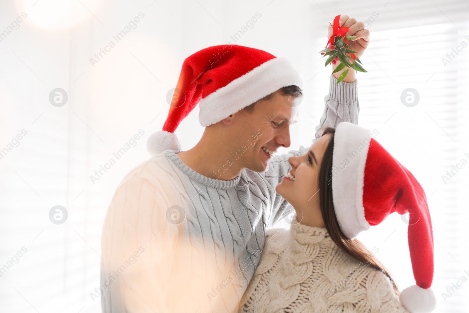 Photo of Happy couple in Santa hats under mistletoe bunch at home, bokeh effect