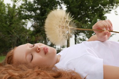 Cute girl with beautiful red hair blowing large dandelion while lying on green grass in park. Allergy free concept