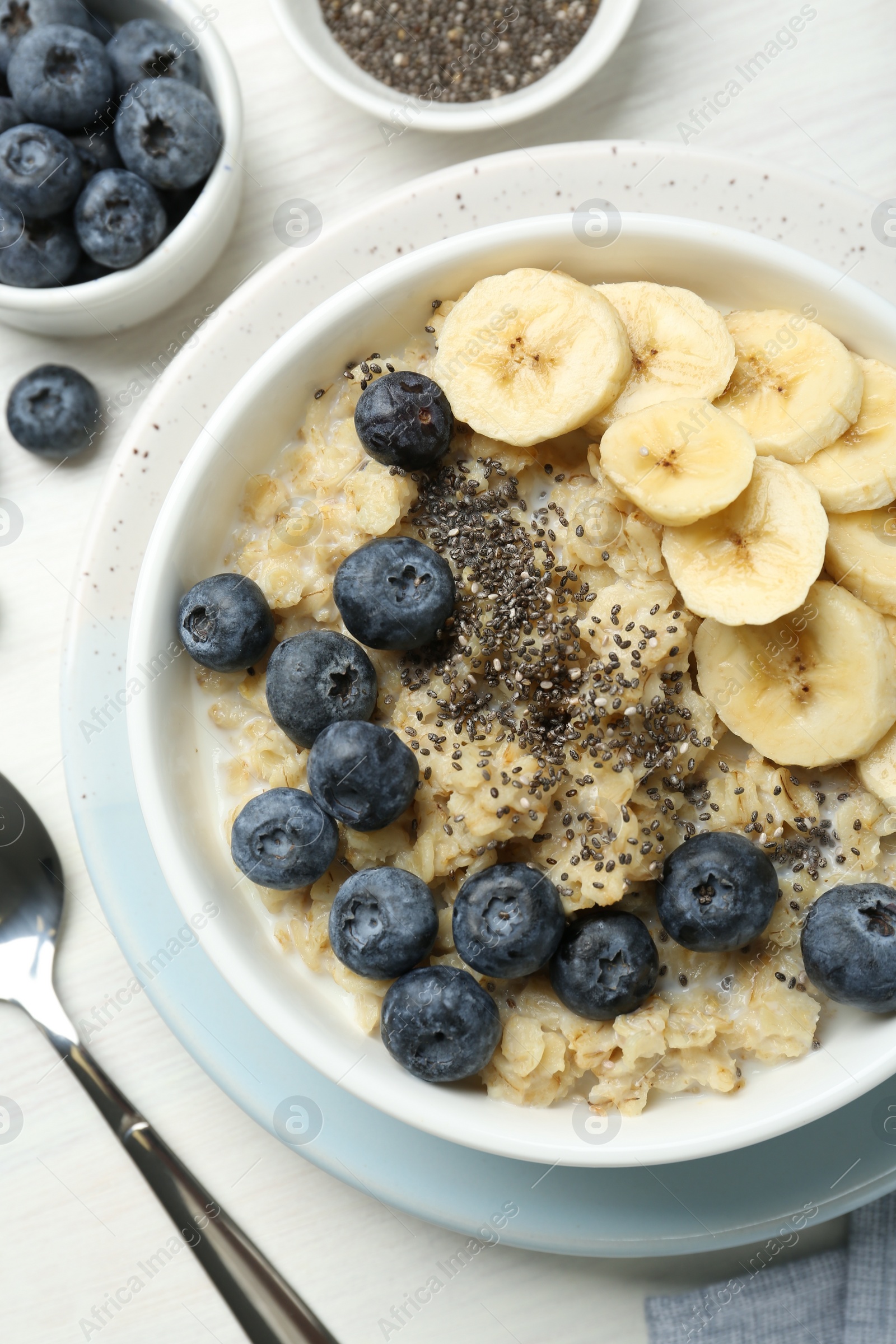 Photo of Tasty oatmeal with banana, blueberries and chia seeds served in bowl on white wooden table, flat lay