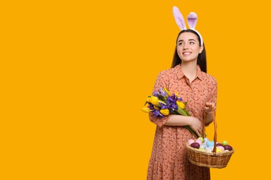 Photo of Happy woman in bunny ears headband holding wicker basket with painted Easter eggs and bouquet of flowers on orange background. Space for text