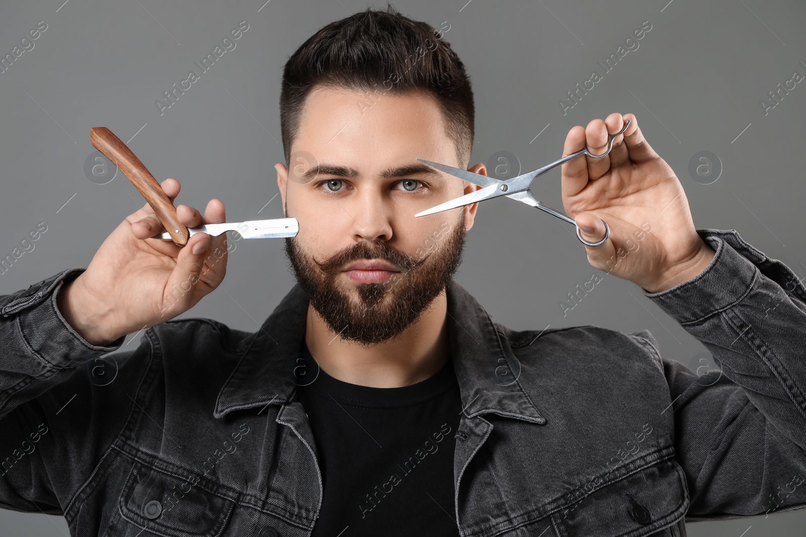 Photo of Handsome young man with mustache holding blade and scissors on grey background