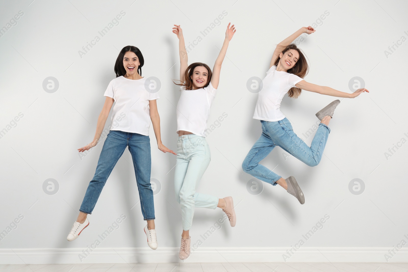 Photo of Group of young women in stylish jeans jumping near light wall