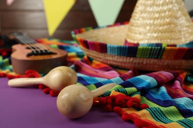 Photo of Mexican sombrero hat, poncho and wooden maracas on purple table, closeup